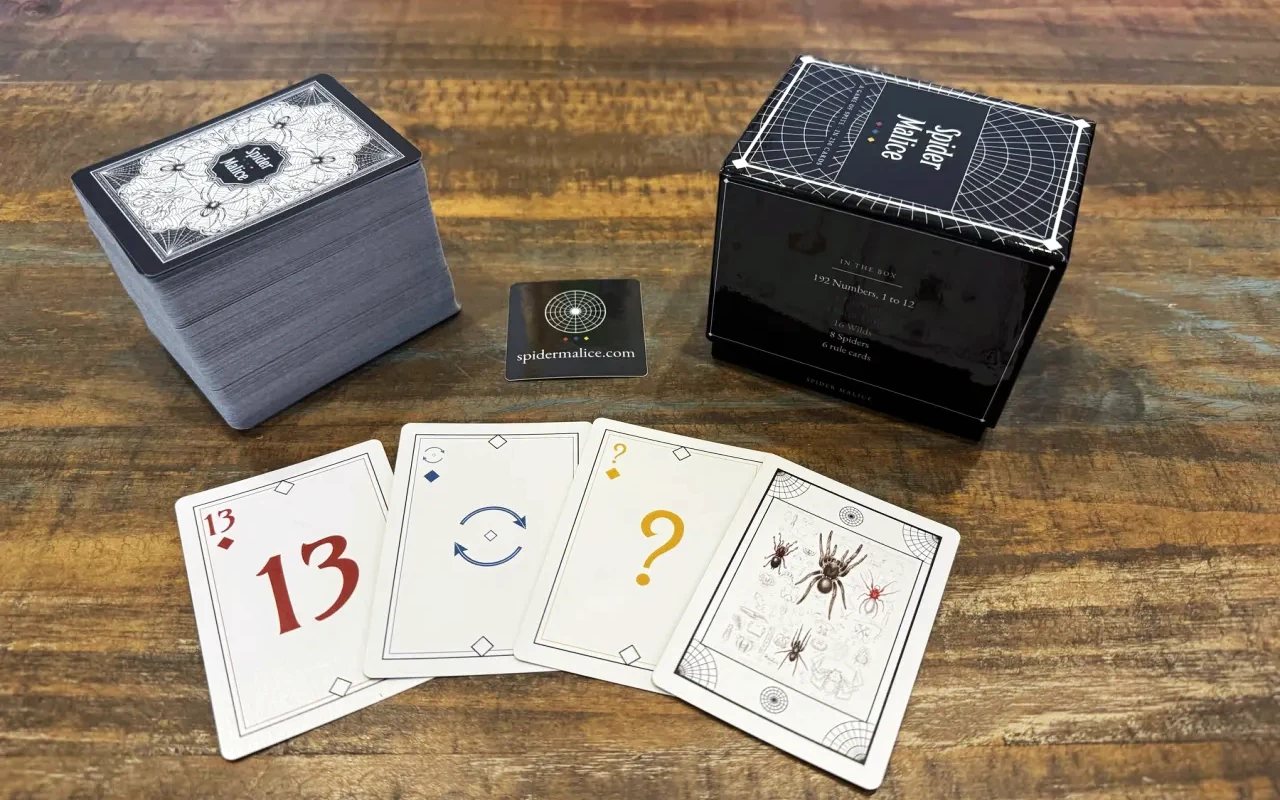 The printed Spider Malice 1.0 deck on a wooden table: the stacked deck showing the engraved back, the black box with its contents list, a spidermalice.com sticker, and a red Thirteen, a blue Extra Turn, a yellow Wild and a Spider fanned in front.